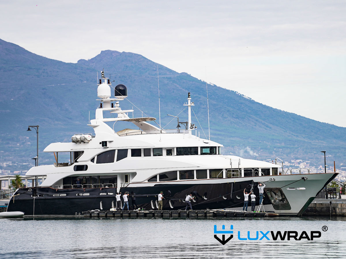 LuxWrap team installing blue marine vinyl film on a superyacht hull from floating rafts